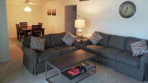 A living room with two grey sofas, patterned cushions, a coffee table, a lamp, a clock, and a dining area with chairs and a table. - 2 Bedroom condo in lovely quiet Naples location close to beach and shops (Naples)