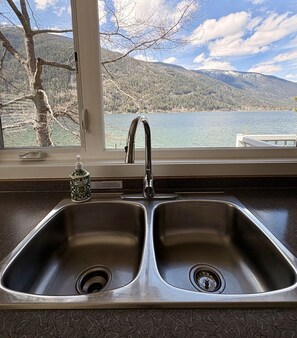 A kitchen with a double stainless steel sink and a window overlooking a lake and mountains.