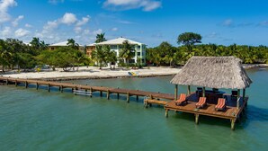 Un resort con una estructura de techo de paja, muelle de madera y un edificio con varios balcones con vista al agua.