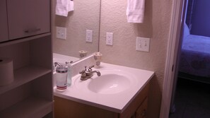 A bathroom with a white sink, a mirror, and a toilet. There are white towels hanging on the wall and a shelf with a roll of toilet paper. - 2/2 Condo, Ground Floor, 2 Parking Spaces, Pool (Saint Augustine Beach)