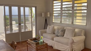 Ein geräumiges Wohnzimmer mit weißem Sofa, einem Esstisch und einer Stehlampe. Große Fenster bieten Blick ins Freie.