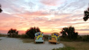 Two beach chairs with a painted design, positioned on a sandy area with grass patches and palm trees in the background, under a sunset sky.