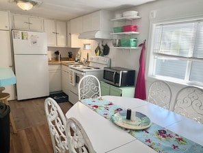 Cocina compacta con armarios blancos, microondas, estufa y una mesa de comedor con mantel floral.