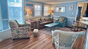 A living room with a sofa, armchairs, a coffee table, and a small stool. There are two lamps, a rug, and artwork on the walls.