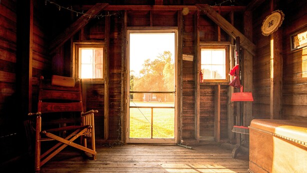 A rustic wooden cabin interior with a large door, a wooden bench, and a small table. - Peaceful Cozy Home In A Rural Garden Farm Setting ~ "Bobolink" House (Carrington)