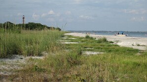 A beach with sand, grass, and a lighthouse in the distance.