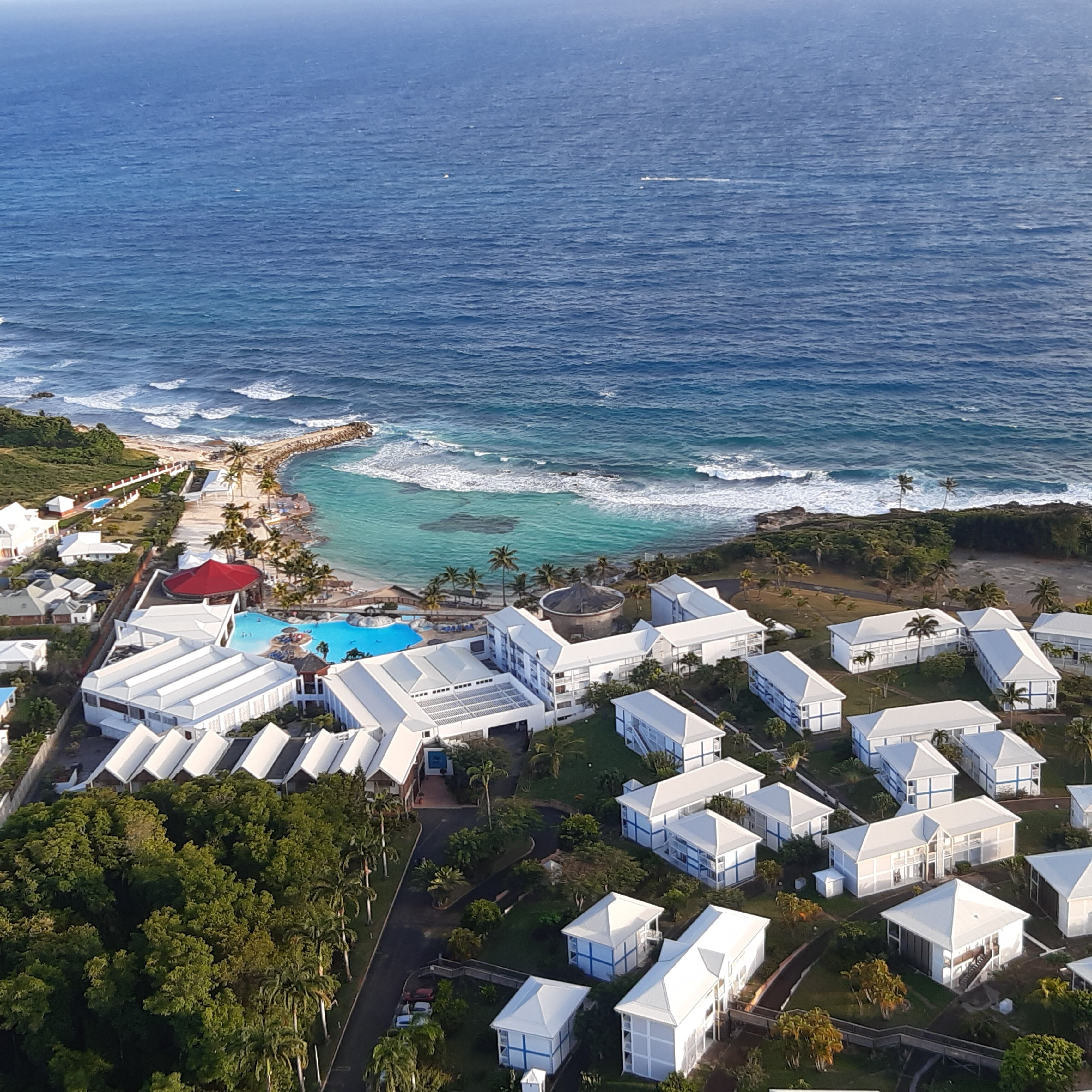 An aerial view of a coastal resort with multiple white buildings, a swimming pool, and a beach area.