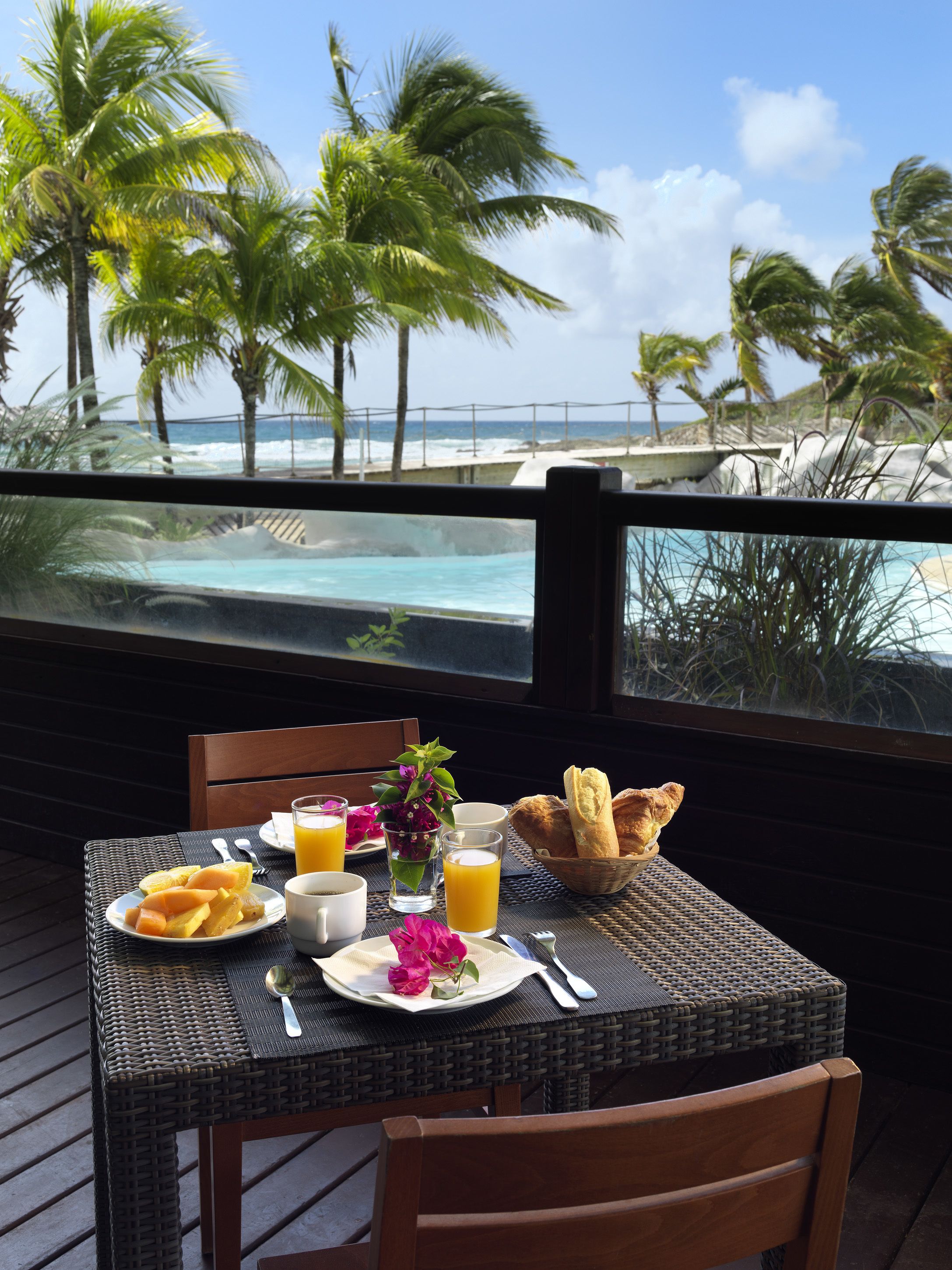 A table set with breakfast on a balcony overlooking a beach with palm trees and ocean waves.