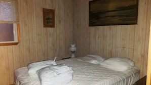A hotel room with wooden panel walls, a bed with white bedding, a framed picture on the wall, and a bedside lamp.