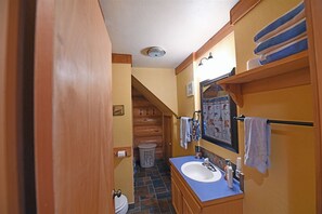Ein kleines Badezimmer mit einem Holz-Waschtisch, einem Waschbecken und einem Spiegel. Ein Handtuchhalter mit Handtüchern, eine Toilettenpapierhalterung und ein Mülleimer sind vorhanden.