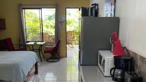 Quarto de hotel com cama, pequena mesa de jantar, cadeiras, cozinha compacta e vista para o verde lá fora.