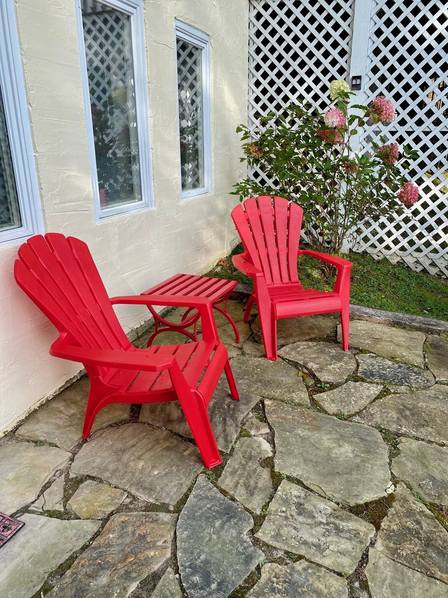 Dos sillas Adirondack rojas en un patio de piedra con una cerca blanca y una casa al fondo.