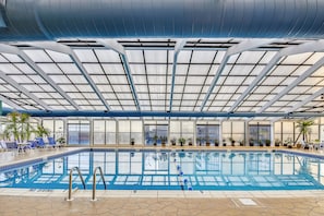 Uma piscina interior com teto de vidro, cadeiras azuis à beira da piscina e plantas em vasos.
