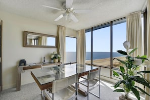 Quarto de hotel com mesa de vidro, ventilador de teto, vista para a praia e uma janela grande.
