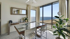 Una habitación con una mesa de vidrio, sillas, un ventilador de techo, un amplio ventanal con vista a la playa y una planta en maceta.