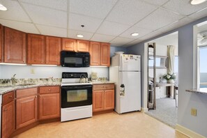 A kitchen with wooden cabinets, a black microwave, a white refrigerator, and a stove. - XPLORIE ACTIVITIES & LINENS INCLUDED*! Capri 2BR/2BA Condo with Ocean & Bay Views! Sunday-Sunday reservations. (Ocean City)