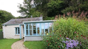 A quaint cottage with a gravel path leading to a wooden door, surrounded by lush greenery and flowering plants. - Tinners Forge - One Bedroom Cottage, Sleeps 2 (Polgooth)