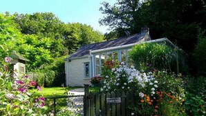 A quaint cottage with a garden entrance, surrounded by lush greenery and flowering plants. - Tinners Forge - One Bedroom Cottage, Sleeps 2 (Polgooth)