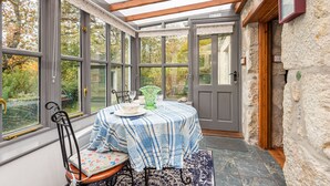 A conservatory with a table set for dining, large windows, and a stone wall. - Tinners Forge - One Bedroom Cottage, Sleeps 2 (Polgooth)