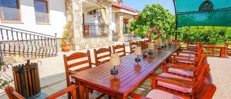 Un espacio de comedor al aire libre con una larga mesa de madera y sillas, un edificio de piedra al fondo y un toldo verde que da sombra.