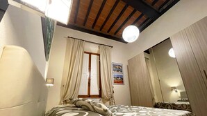 Ein Schlafzimmer mit Bett, Vorhängen, einem Fenster, einem Spiegel und einer Deckenleuchte.