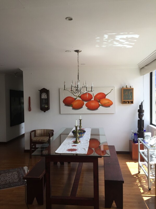 A modern living room with a glass-top dining table, a wooden bench, a chandelier, a clock, and a large fruit-themed painting. - Beautiful, safe, centrally located; add private driver with car for $40per day (Quito)