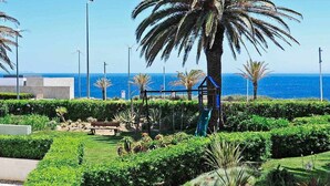 Un jardín ajardinado con palmeras, un parque infantil y vista al océano.