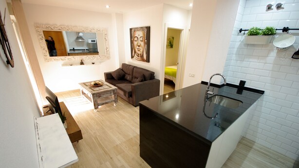 Eine moderne Küche mit schwarzem Arbeitsplatz und Spüle. Es gibt eine Essecke mit Tisch und Stühlen, sowie ein Sofa. Die Wände sind weiß und ein Buddha-Gemälde ist an der Wand angebracht.