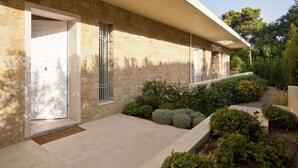 Een stenen gebouw met een witte deur, een kleine tuin met diverse planten, en een heldere lucht.