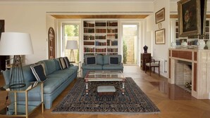 Een woonkamer met een blauwe bank, een glazen salontafel, een vloerkleed met een patroon en een boekenkast vol boeken.
