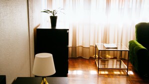 Una stanza con pavimento in legno, un tavolino nero con una lampada, una poltrona verde, un piccolo tavolo con un libro e una pianta in vaso su un mobile nero.