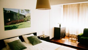 Una camera d'albergo con un letto, un comodino, una lampada, una cassettiera, una sedia e una finestra con tende.