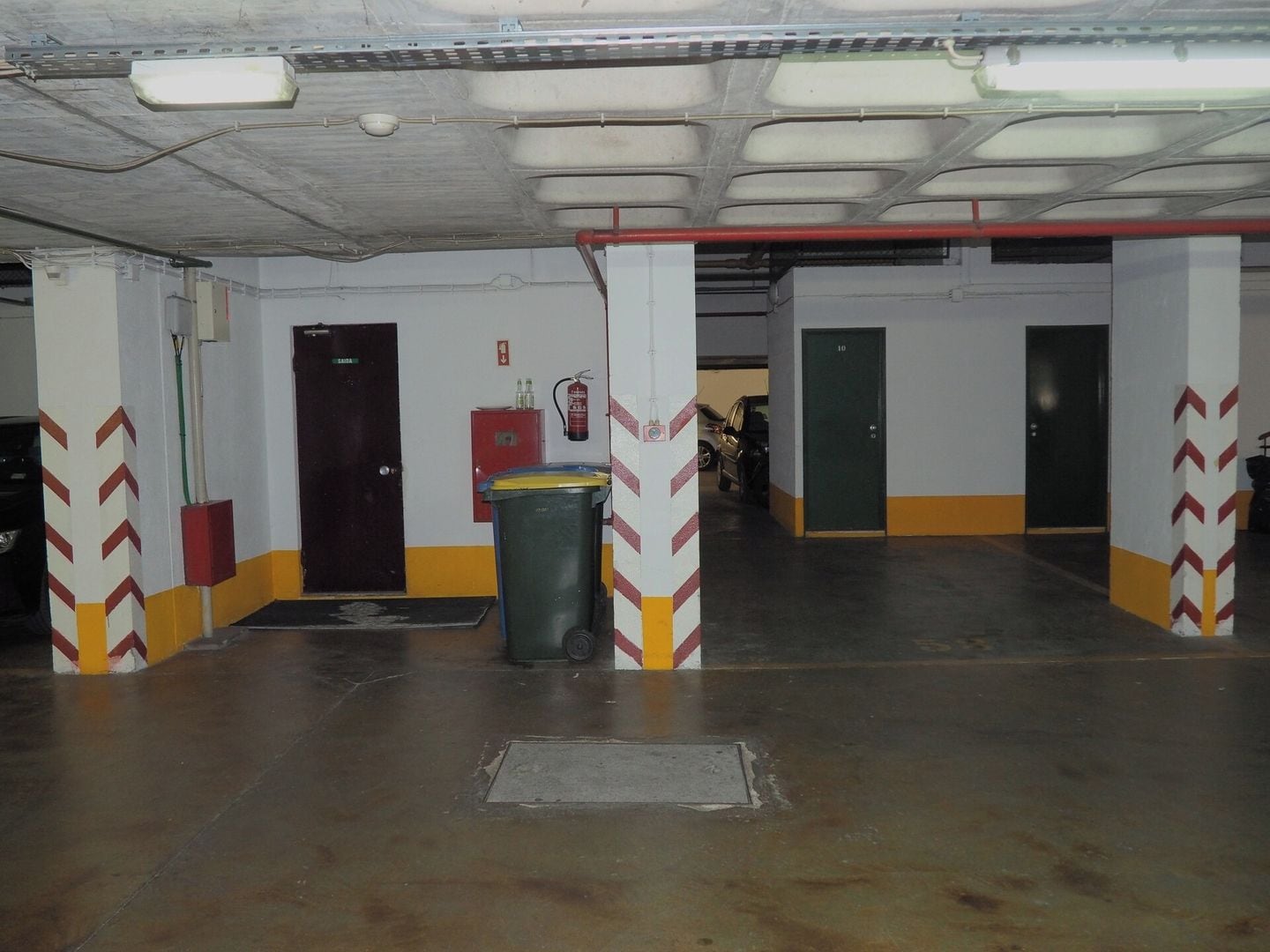 A parking garage with concrete pillars, a trash bin, and doors leading to other areas.