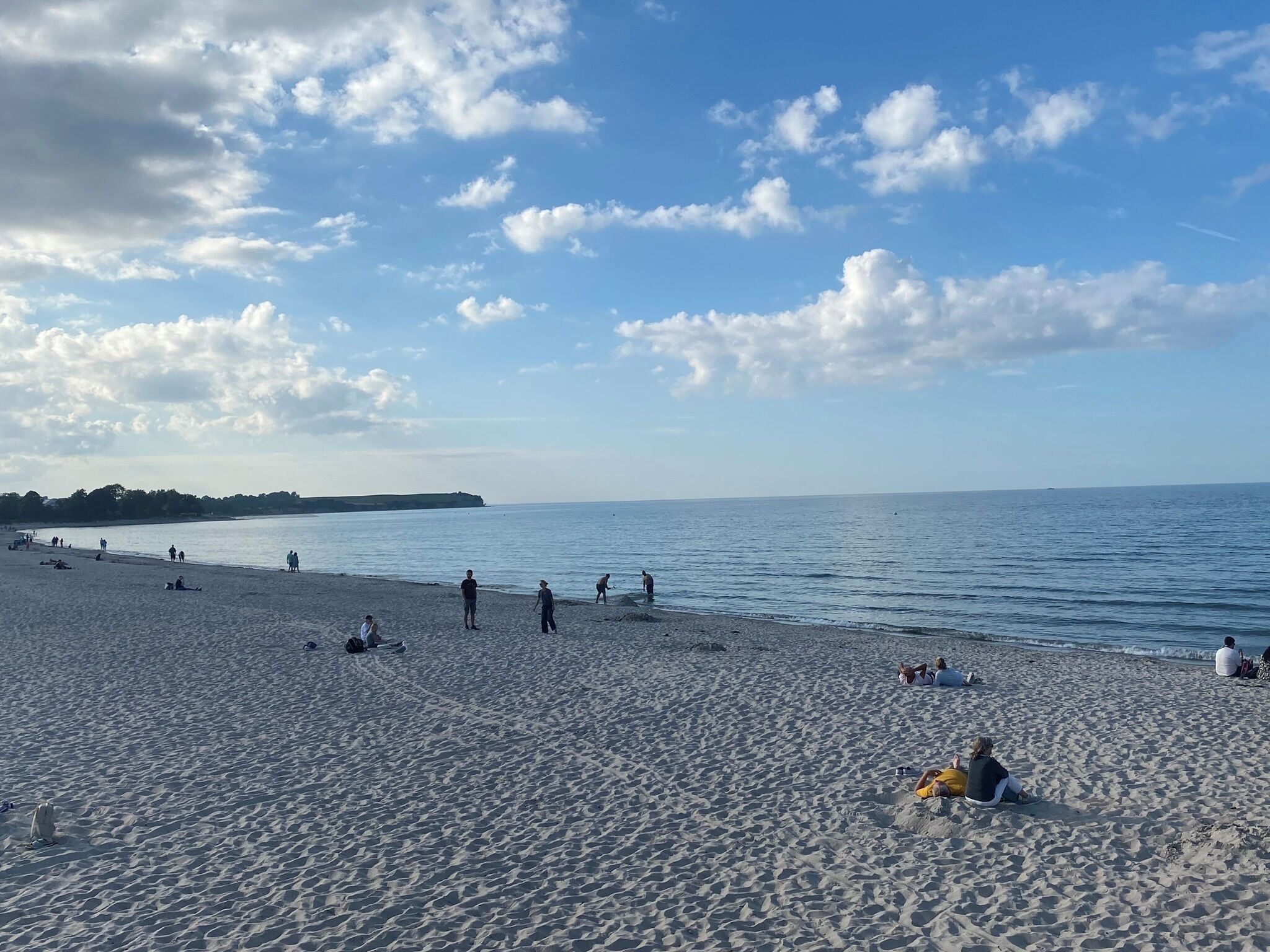 Ein Sandstrand mit Menschen, die entlanggehen und sitzen, ruhiges Meer und teilweise bewölkter Himmel.