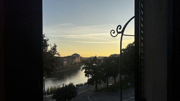 A view from a window overlooking a river, a pedestrian crossing, and a curved road with trees and buildings in the background. - Rivadige Suite 3 - Verona Journeys (Verona)