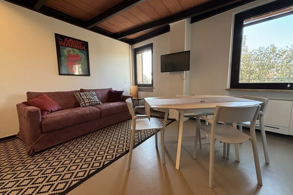 Una sala de estar con un sofá marrón, un tapete estampado, una mesa blanca con sillas, un televisor en la pared y una ventana con vistas.