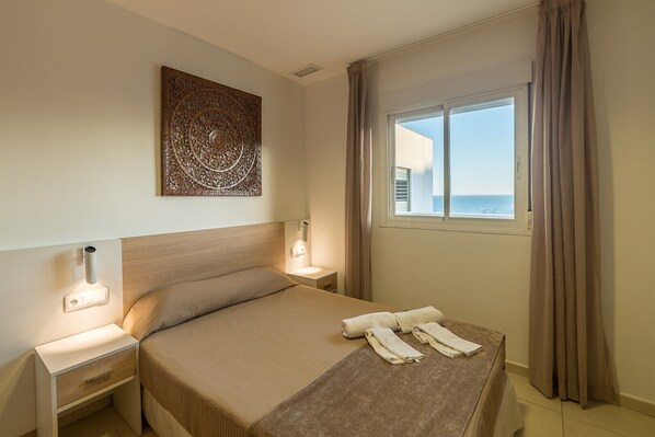 A hotel room with a bed, a nightstand, a wall-mounted artwork, a window with curtains, and a view of the sea.