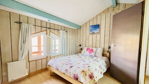 Quarto com paredes de madeira, uma cama com roupa de cama florida, uma janela com cortinas, um radiador e uma porta.