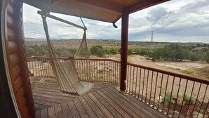 Una terraza de madera con una silla mecedora, barandilla y vistas a un paisaje desértico con vegetación escasa y una torre de líneas eléctricas.