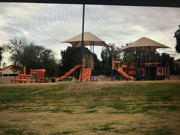A playground with slides and climbing structures, shaded by large umbrellas, and surrounded by grass and trees.