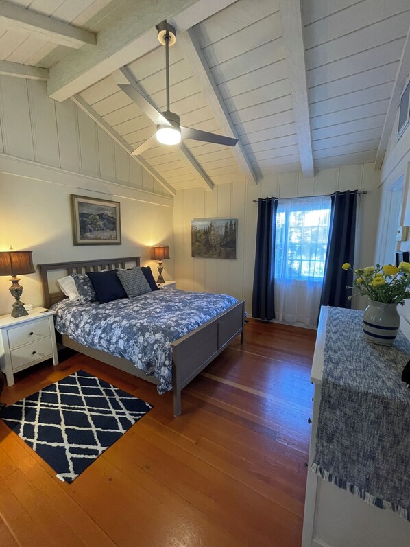 A bedroom with a bed, nightstands, a ceiling fan, a window with curtains, and a small table with a vase.