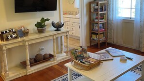 Una sala de estar con televisor, una mesita consola con adornos, una estantería y un ventanal con cortinas.