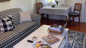 Una sala de estar acogedora con un sofá estampado, una mesa de centro con revistas, una mesa de comedor con mantel floreado y una ventana con cortinas traslúcidas.