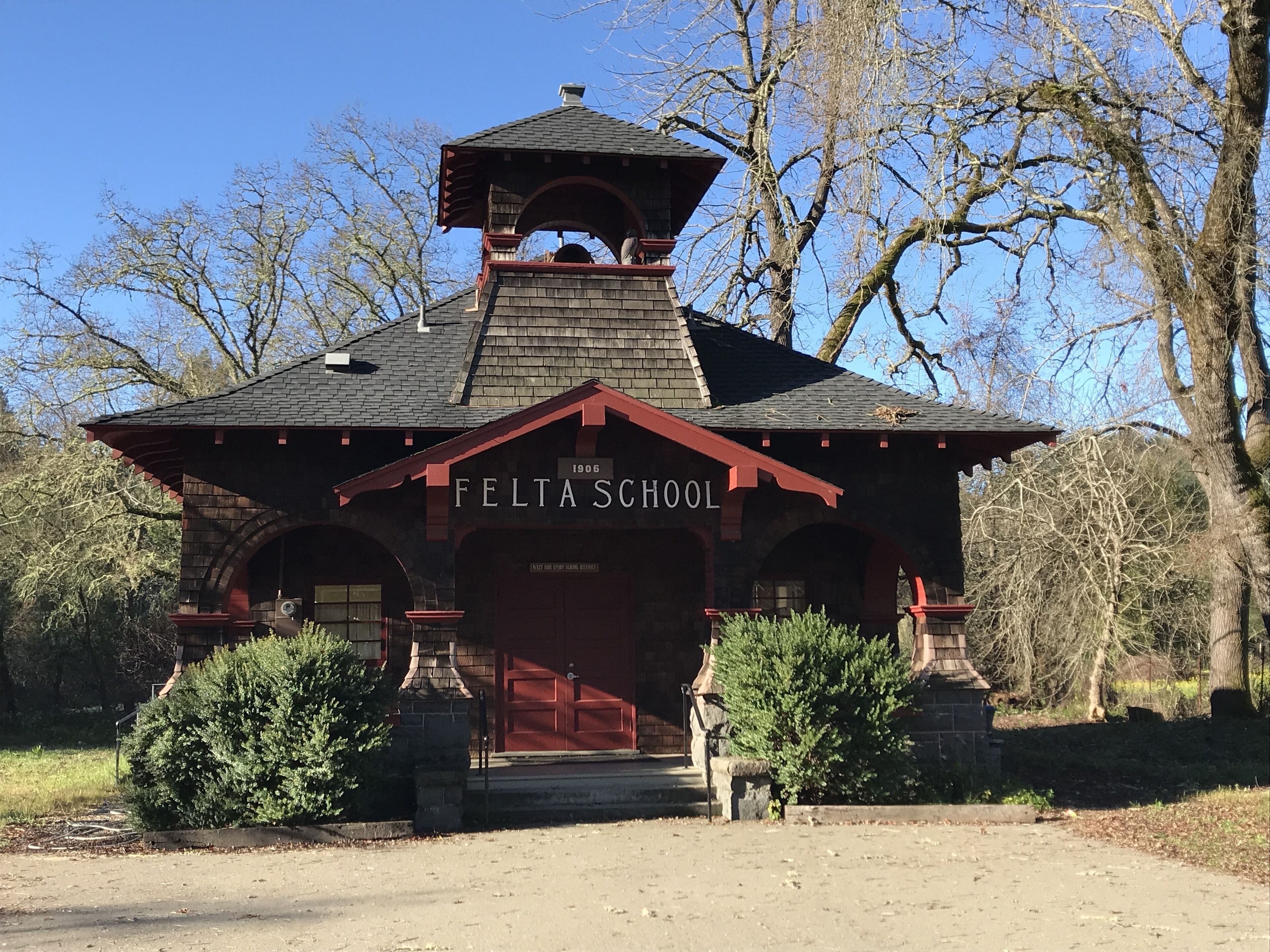 A historic school building with a red roof and arched entrance, surrounded by trees and a clear sky.