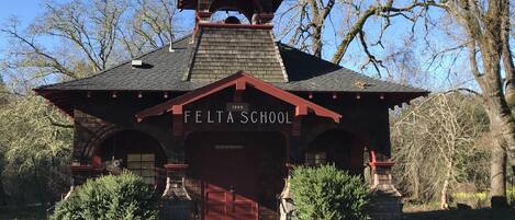 A historic school building with a red roof and arched entrance, surrounded by trees and a clear sky.