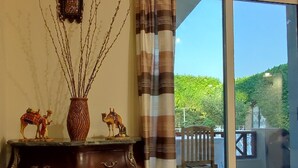 Ein Zimmer mit einem hölzernen Schminktisch, Dekorationen, Blick auf einen Balkon mit Grünpflanzen und einer Glastür.