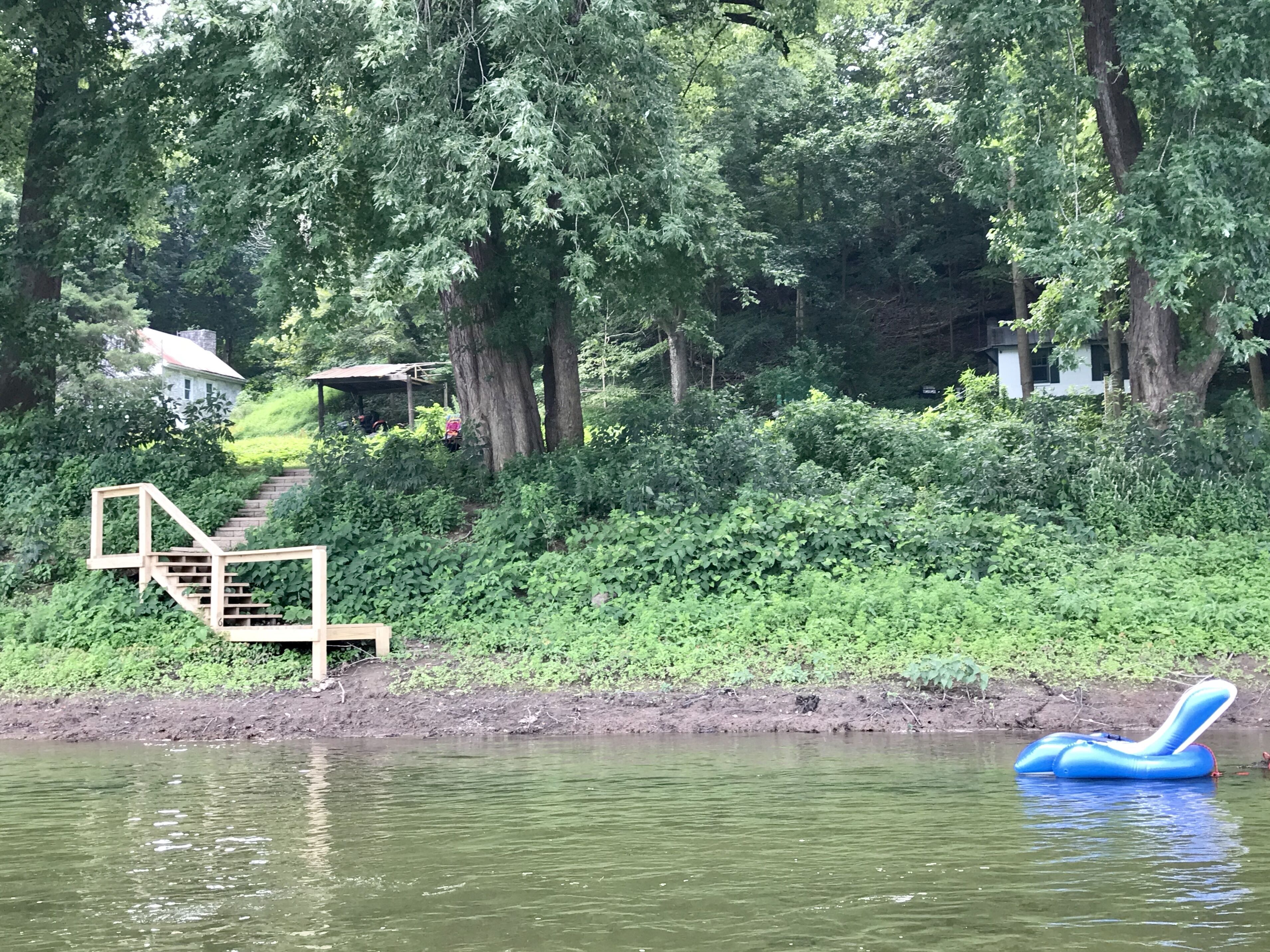 A serene lakeside setting with a wooden staircase leading to a covered area, surrounded by lush greenery and trees.