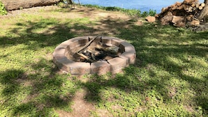 A circular fire pit surrounded by grass and small plants, with a bench and logs nearby. - ❤️ Secluded 1940s Romantic Tiny House on the River (Shepherdstown)