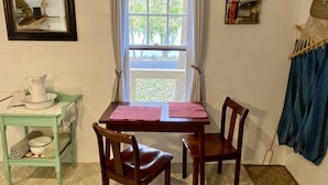 A room with a wooden table, two chairs, a small shelf with a pitcher and bowl, a framed picture, and a window with curtains. - ❤️ Secluded 1940s Romantic Tiny House on the River (Shepherdstown)