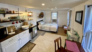 A compact kitchen with white cabinets, a sink, and a countertop with various kitchen items. There is a bed with a patterned bedspread and pillows, and a window with curtains. - ❤️ Secluded 1940s Romantic Tiny House on the River (Shepherdstown)